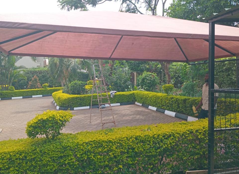 Brown shade net carport roof over a paved parking area in a landscaped garden