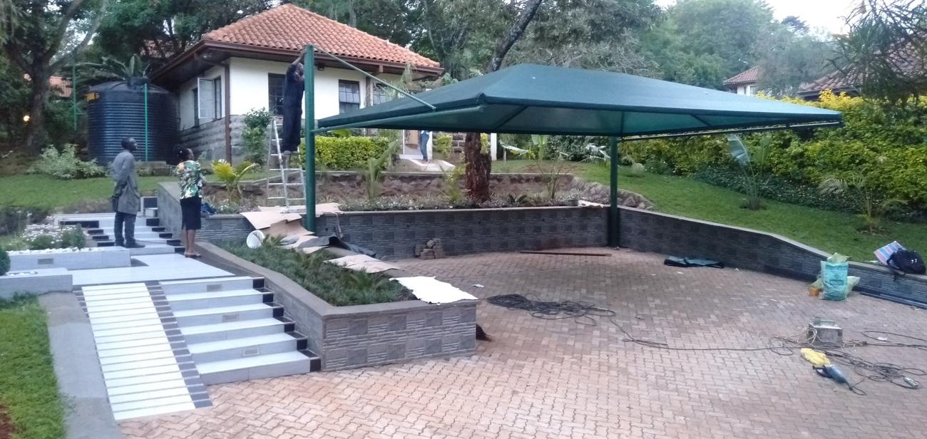 Green shade net carport on steel poles being installed over a paved yard beside a garden